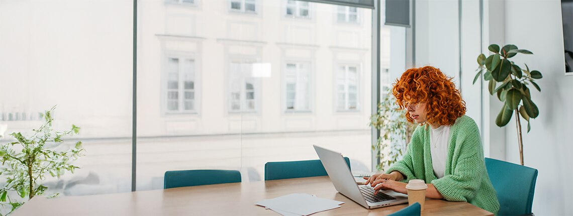 A woman sitting at a desk by the window on her laptop.
